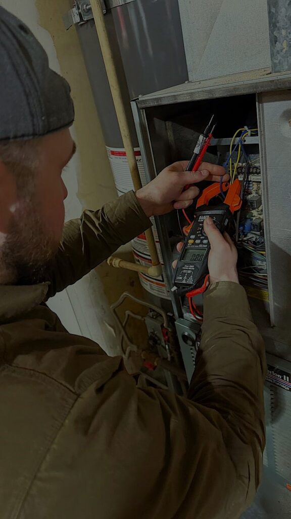 HVAC repairman using a digital multimeter to test the electrical control board of a residential heating system during a diagnostic visit.
