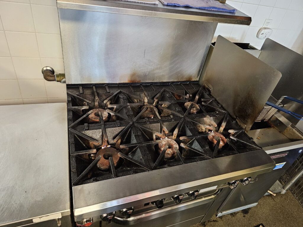 Top-down view of a 6-burner commercial gas stove top in a Seattle restaurant featuring heavy-duty cast iron grates and high-BTU burner heads before professional cleaning.