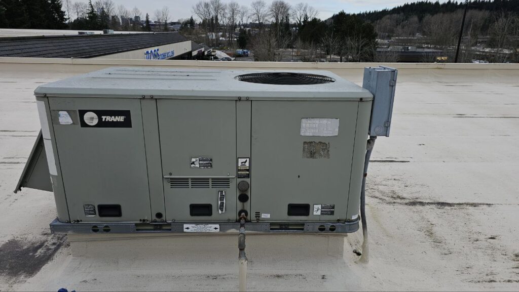 A large grey Trane commercial rooftop HVAC unit sitting on a white membrane roof. The unit features several access panels and an electrical disconnect box mounted on the side.