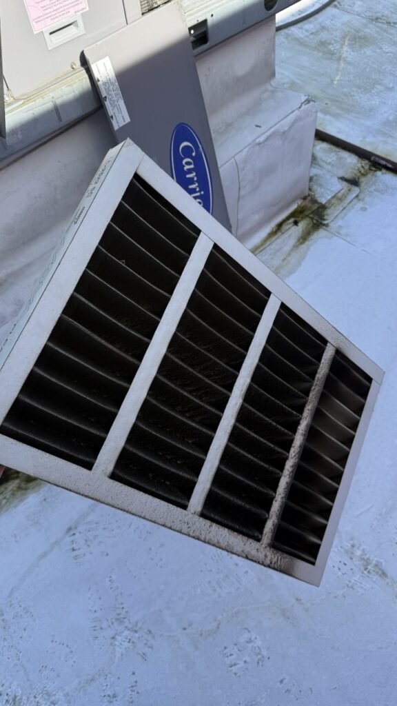 A/C Dr. Naz technician holding a dirty commercial air filter next to a Carrier rooftop unit during a preventive maintenance visit in Seattle.