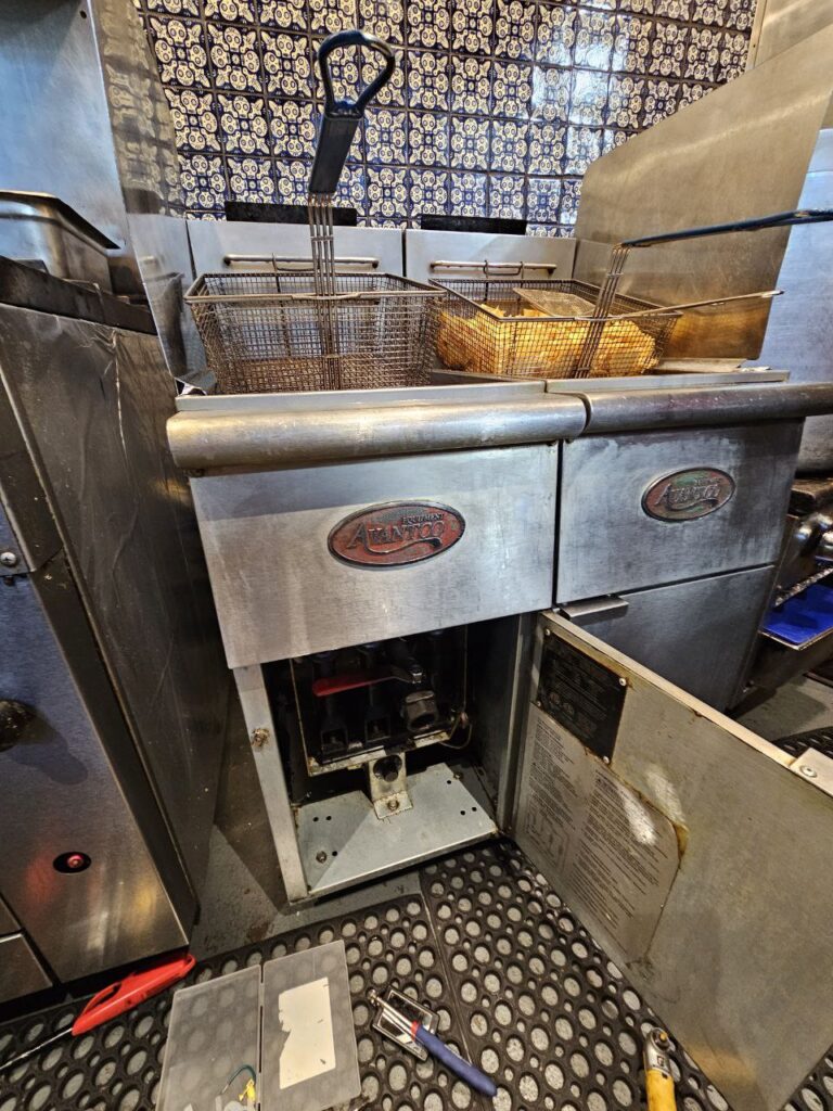 On-site repair of an Avantco double-tank commercial deep fryer in a restaurant kitchen, with the lower service door open to reveal the burner compartment and gas manifold.