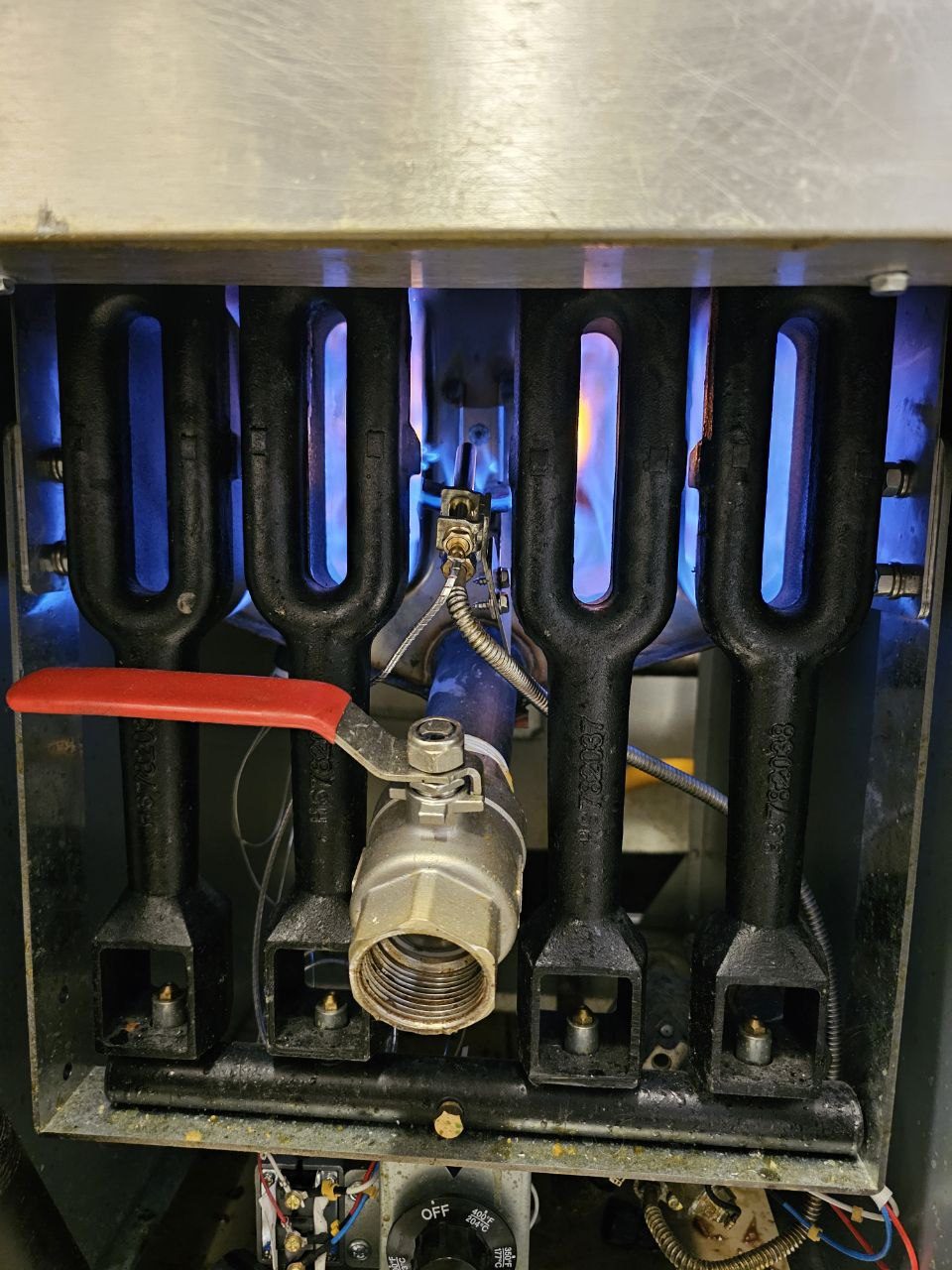 A close-up view of the internal burner assembly of a commercial gas fryer featuring four black cast-iron burners with visible blue flames and a central gas valve with a red handle.