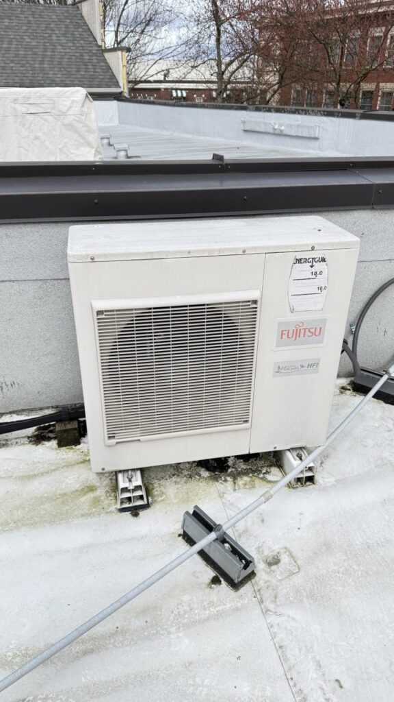 A Fujitsu Halcyon HFI commercial heat pump unit requiring repair on a Seattle commercial rooftop, positioned near a parapet wall for facility climate control.