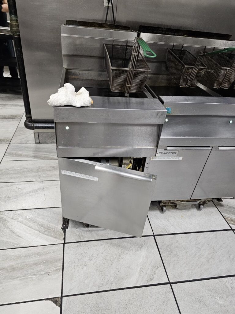 A close-up view of a commercial fryer's gas manifold and burner system, featuring a stainless steel ball valve with a red handle and four cast-iron burner tubes ignited with consistent blue flames.