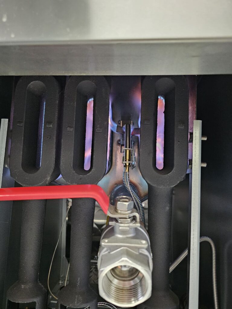 An interior view of commercial fryer heat tubes and burner assembly with a red-handled gas valve and a lit pilot flame, demonstrating the proper ignition sequence for gas-powered kitchen equipment.