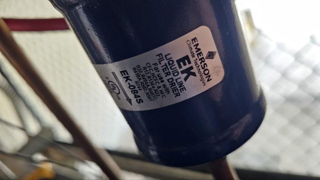 Close-up of an Emerson EK Liquid Line Filter Drier (Model EK-084S) installed on a copper refrigerant line, used to remove moisture and contaminants from the system.