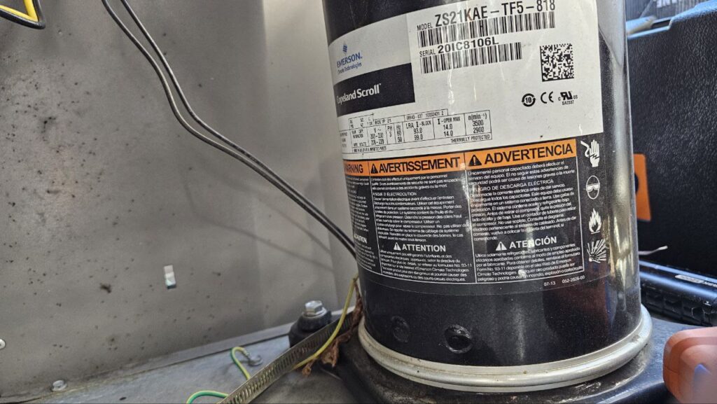 Close-up of a black Copeland Scroll compressor (Model ZS21KAE-TF5-818) inside a commercial AC unit. The label shows technical specifications and safety warnings.