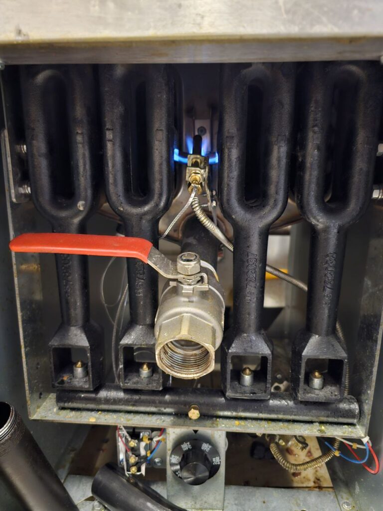 A close-up, front-facing view of a commercial fryer's burner manifold, featuring four heavy-duty cast-iron burners, a stainless steel gas shut-off valve with a red handle, and a lit pilot light positioned centrally to ensure proper ignition.