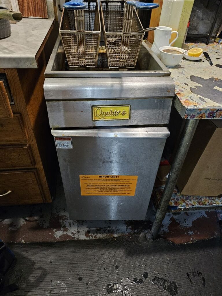 A Qualite brand commercial floor fryer with blue-handled baskets and a prominent warning label regarding proper operation, positioned in a professional cook line.