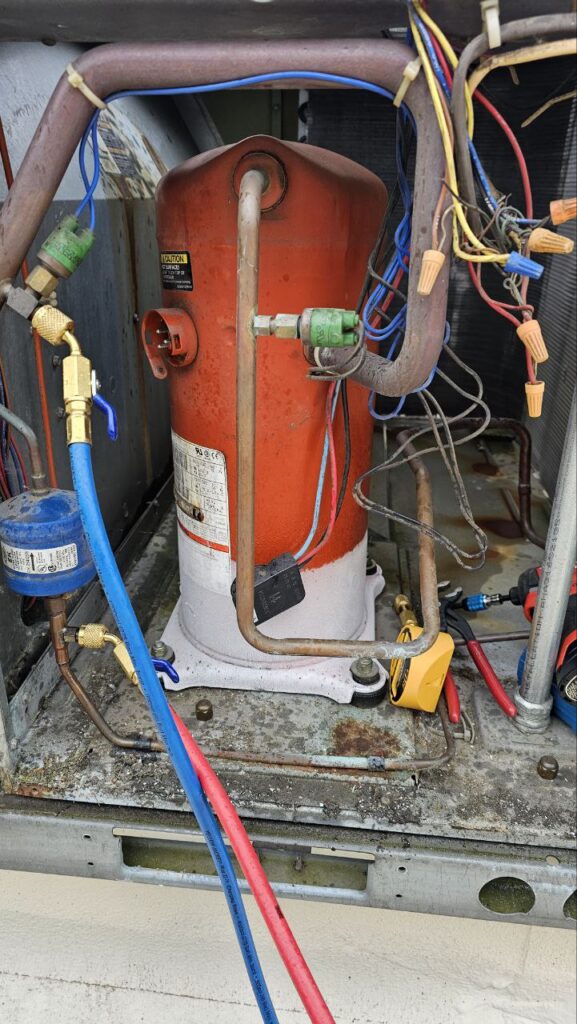 The interior of an HVAC cabinet showing an orange Copeland compressor with ice or heavy frost buildup at the base. Service hoses are connected to the ports, and various electrical wires are visible.
