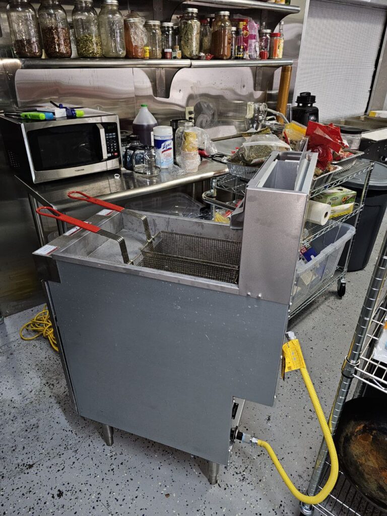 A portable commercial floor fryer on heavy-duty casters, showing the open cabinet base with the gas control valve and pilot light assembly ready for a mobile service repair.