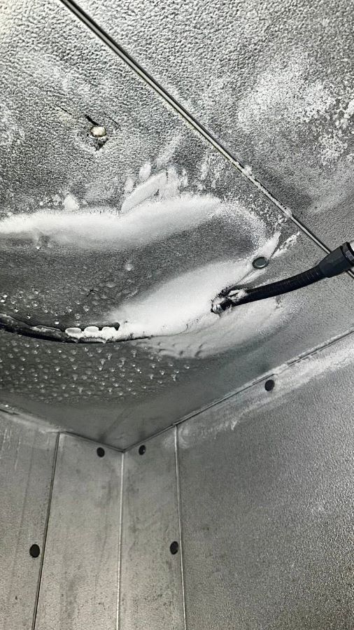 Walk-in freezer ceiling corner with heavy frost and ice buildup around a conduit penetration on stippled metal panels.