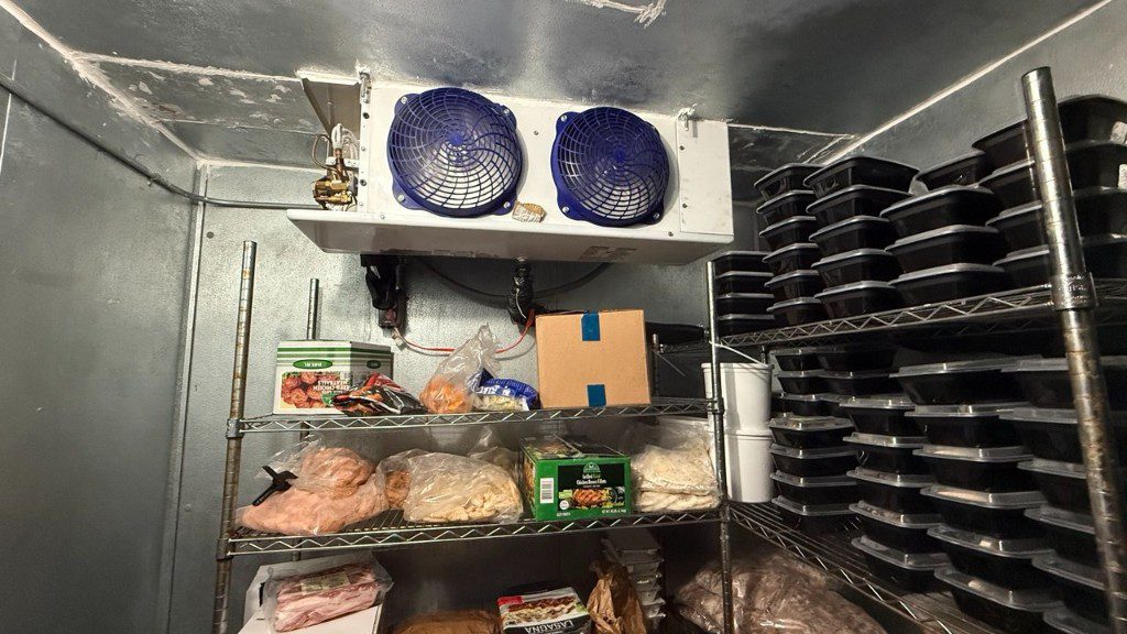 Commercial walk-in cooler stocked with food and a ceiling-mounted evaporator with blue fan guards